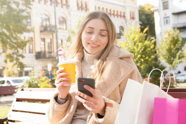 公園の外にコーヒーを持っている陽気な女性 彼女のスマートフォンを使用して — ストック写真