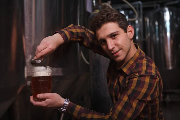 タンクから醸造されたばかりのビールを彼のマイクロブルワリーでマグカップに注ぎながら カメラを見ている陽気な醸造者 — ストック写真