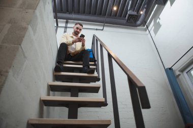 Akıllı telefon kullanan yakışıklı bir adamın düşük açılı görüntüsü. Sırt çantasıyla modern otel odasındaki merdivenin tepesinde oturuyor.