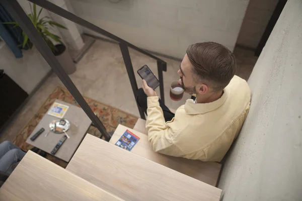 階段の屋内に座って 彼のスマートフォンにテキストメッセージをしながら 男がお茶を持っているのトップビューショット — ストック写真
