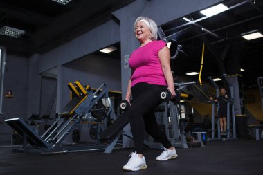 Neşeli yaşlı kadın gülüyor, dambıllarla hamle yapıyor.