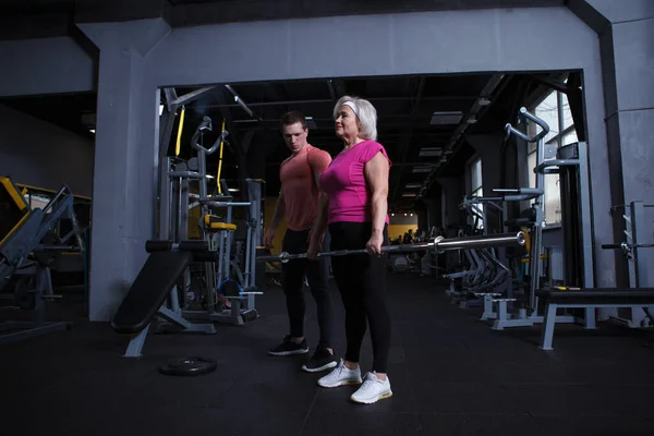 Full length shot of an elderly woman lifting barbell, working out with personal trainer at the gym