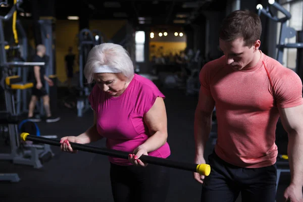 Healthy senior woman exercising with personal trainer, doing bicep curls with bodybar
