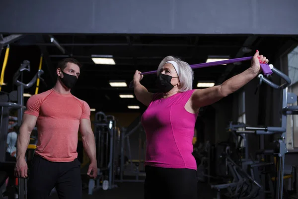 Elderly woman stretching at the gym with resistance band, wearing medical face mask