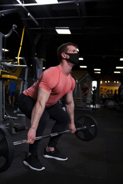 体操で重い鐘を持ち上げ 医療面マスクを身に着けている筋肉スポーツマンの垂直ショット — ストック写真