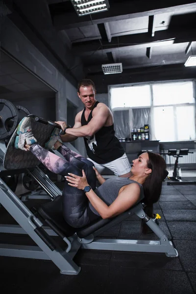 Vertical shot of a professional personal trainer assisting his female client in leg press gym machine workout