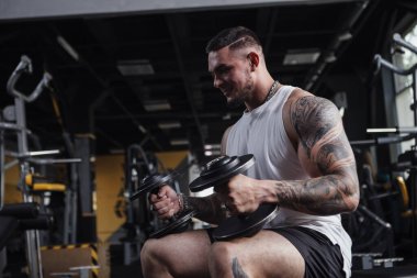 Handsome tattooed muscular bodybuilder holding heavy dumbbells, sitting at the gym