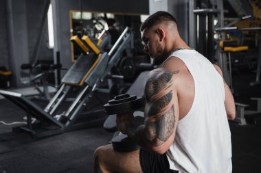 Rear view shot of a big muscular man resting at gym, holding heavy dumbbells, copy space