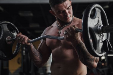 Hndsome tattooed muscular sportsman exercising shirtless at gym, doing barbell biceps curls