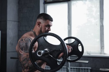 Huge tattooed muscular man working out with barbell, doing biceps curls