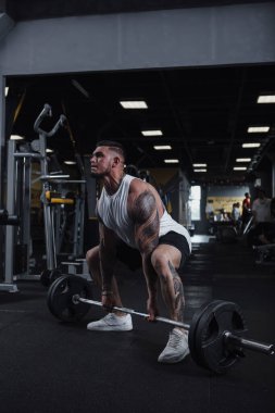 Vertical full length shot of a strong muscular tattooed male athlete working out at fumctional training gym, lifting barbell