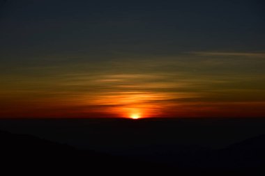 Güneş sık bir ormanın arkasında batıyor ve ardından dağlar geliyor.