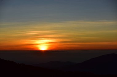 Güneş sık bir ormanın arkasında batıyor ve ardından dağlar geliyor.