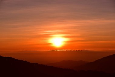 Güneş sık bir ormanın arkasında batıyor ve ardından dağlar geliyor.