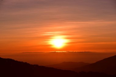 Güneş sık bir ormanın arkasında batıyor ve ardından dağlar geliyor.