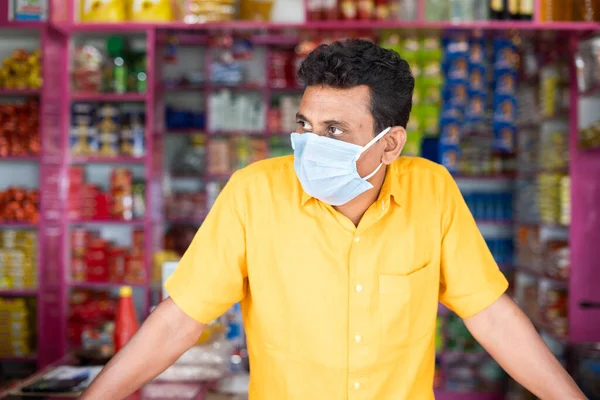 Merchant with mdical face mask at groceries or Kirana store waiting for customers by looking around during coronavirus covid-19 pandemic - concept of small businesses reopen and new normal lifestyle.