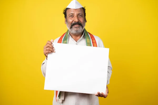 Portrait d'un politicien indien souriant heureux tenant un panneau blanc vide en regardant la caméra sur fond de studio jaune. — Photo