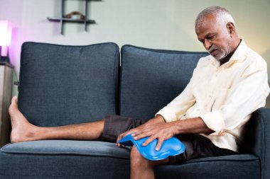 Son sınıf öğrencisi evde koltukta otururken diz eklem ağrısı için sıcak su torbası kullanıyor. Doğal sıcak su tedavisi, kas gevşemesi ve osteoartrit kavramı..