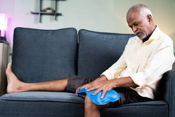 Senior man using Hot water massage bag for knee joint pain relief while sitting on sofa at home - concept of natural hot water treatment, muscle relaxtion and osteoarthritis. — стоковое фото