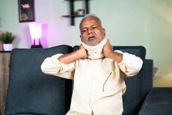 Indian senior old man using neck brace or carvical collars due to neck pain at home - concept of arthritis and neck pain relief treatment. — стоковое фото