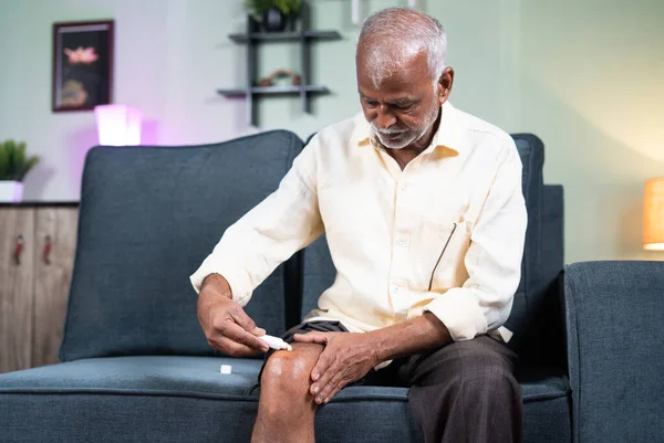 Focus on hands, Senior man applying ointment cream for joint knee pain at home while sitting on sofa - έννοια της θεραπείας για οστεοαρθρίτιδα, τραυματισμό στο γόνατο και αρθρίτιδα — Φωτογραφία Αρχείου