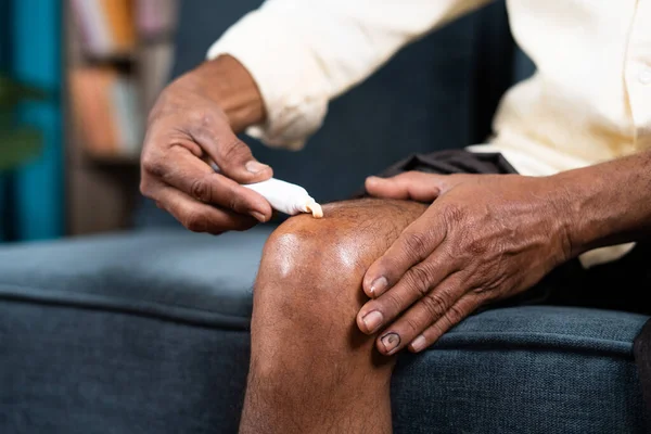 Primer plano del anciano irreconocible que aplica crema ungüento para el dolor de rodilla articular en el hogar - concepto de tratamiento o terapia para la osteoartritis y el esguince de rodilla — Foto de Stock