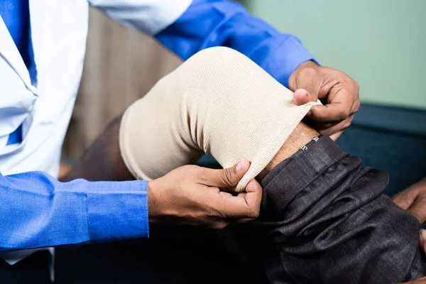 Close up shot of unrecognizable Physician treating old man by wearing stretch bandage for knee sprain or joint paint- concept of senior people muscle injury, healthcare treatment. — стоковое фото