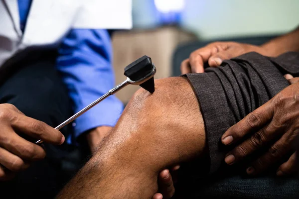 Close up shot of unrecognizable physician checking knee joint pain of senior man by using knee hammer at home - concept of healthcare, treatment for injury and wellness. — стоковое фото