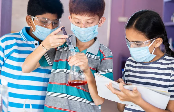 Diversos niños que experimentan con anteojos de seguridad y mascarilla facial observando los resultados mientras mezclan productos químicos usando frasco en el laboratorio de química - concepto de educación, aprendizaje y desarrollo infantil. —  Fotos de Stock