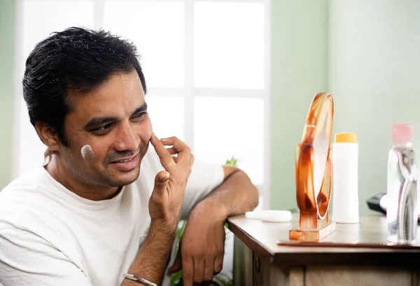 Beau jeune homme appliquant une crème anti-âge ou hydratante sur le visage en regardant miroir à la maison concept de protection des soins de la peau, problème de peau et d'autosoin. — Photo
