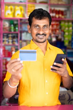 Kirana or groceries merchant showing credit card to camera after on mobile phone - concept of accepting Digital payment in retail business, cashless purchase and e-payment using smartphone.