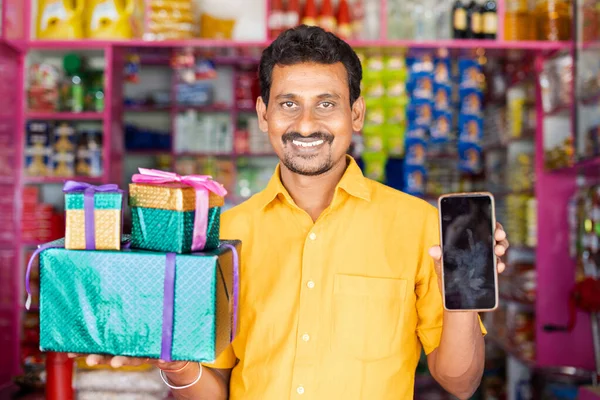 Kirana Shop Merchant with Gifts boxs and mobile phone in hand loking at camera - concpet of Festival sales, promotional offers to win prizes from smartphone. — Photo