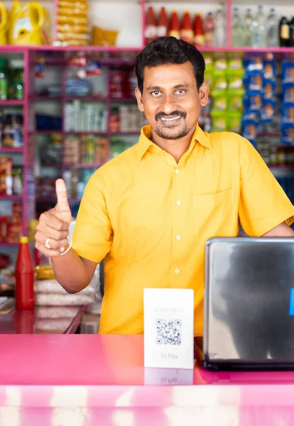 Portrait shot of Kirana or groceries store merchant showing thumbs up sign by looking at camera - concept of successful small business owner, happiness and accepting digital payemnts