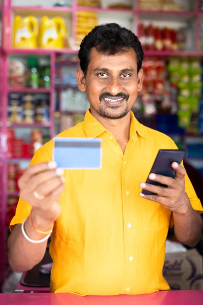 Kirana or groceries merchant showing credit card to camera after on mobile phone - concept of accepting Digital payment in retail business, cashless purchase and e-payment using smartphone. — Photo
