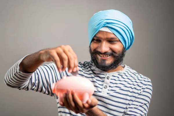 Young indian sikh man saving money by placing coin on piggy bank at grey background - concept of saving and future financial planning and investment. — Fotografia de Stock