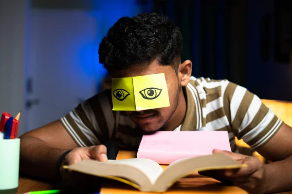 Young student acting like reading while sleeping by putting eye open stickers at home - concept of lazyiness and late night studying, careless and negligence. — Stock Photo, Image