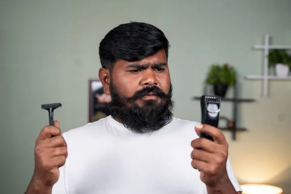young beard man confused or thinking about what to use between razor blade and trimmer or shaver machine at home - concept of grooming, self care and cosmetics