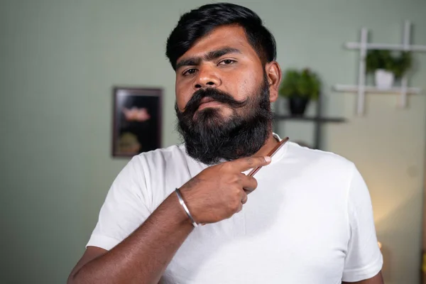 Jeune homme peignant la barbe en regardant la caméra à la maison - concept de soins capillaires, toilettage et bien-être — Photo