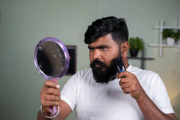 Jeune homme barbe indienne en utilisant un rasoir électrique ou une machine de trimmere en regardant miroir- concept de soins des cheveux, toilettage et bien-être. — Photo