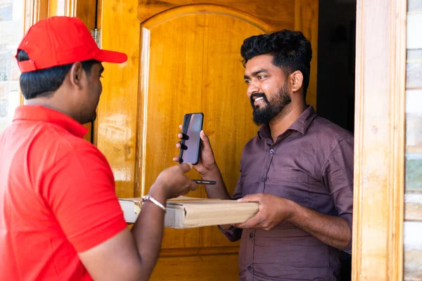 Молодой индийский клиент показывает мобильный телефон после оплаты доставщику, сканируя qr код на мобильном телефоне на пороге - концепция бесценной или безналичной оплаты, технологии и отгрузки. — стоковое фото