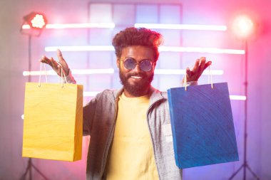 Sahnede, neon arka plandaki kameraya bakarak alışveriş torbalarını gösteren mutlu, şık bir genç adam. Kıyafet reklamları, tanıtım teklifleri ve festival alışveriş satışları kavramı.