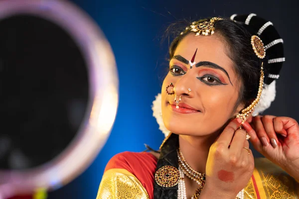 Indiase bharatanatyam danser klaar te maken door het dragen van oorbellen door te kijken naar spiegel - conept van voorbereiding voor dansvoorstelling, traditionele kleding en indiaanse cultuur. — Stockfoto