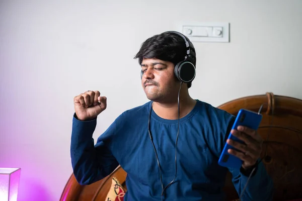 Junger Mann genießt Musik über Kopfhörer, während er im Schlafzimmer Lieder auf dem Handy spielt - Konzept der Unterhaltung, Entspannung und des Nachtlebens — Stockfoto