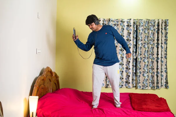 Profiter du jeune homme avec casque dansant en chantant sur le lit en écoutant des chansons sur téléphone portable dans la chambre concept de divertissement, d'activité de loisirs et de détente. — Photo