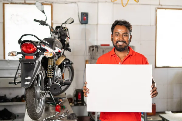 Meccanico di moto sorridente in piedi tenendo vuoto bordo di zucchero bianco o cartello al garage - concetto di promozione aziendale, pubblicità per manutenzione o servizio — Foto Stock