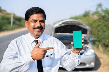 Bozuk arabanın önünde yeşil ekran cep telefonu gösteren mutlu işadamı kameraya bakarak - çevrimiçi mobil araba tamiri, uygulama reklamı ve promosyonu kavramı