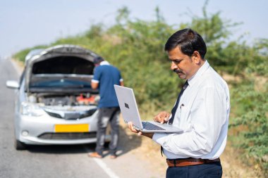 Tamirci araba satın alırken yol kenarındaki işadamı bozuk bir arabanın önünde dizüstü bilgisayarla çalışmakla meşgul - mobil araba servisi kavramı, yoğun çalışma programı ve talihsizlik
