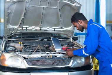 Otomobil tamircisi garajdaki motor veya devreyi kontrol ederken dizüstü bilgisayar kullanıyor - profesyonel, teknoloji, uzmanlık ve bakım hizmeti kavramı.