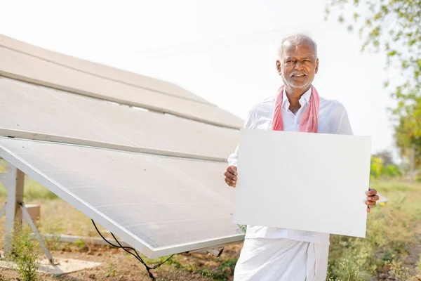 Gülümseyen köy çiftçisi tarım arazisindeki güneş panelinin önündeki kameraya bakarak beyaz tahtayı gösteriyor - tanıtım, reklam ve yenilenebilir enerji kavramı. — Stok fotoğraf