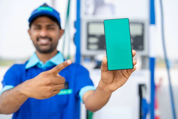 Εστίαση στο τηλέφωνο, χαμογελώντας εργαζόμενος αντλία βενζίνης δείχνει πράσινο οθόνη κινητό τηλέφωνο δείχνοντας δάχτυλο κοιτάζοντας κάμερα στο βενζινάδικο - έννοια της online κράτησης, διαφήμιση και βενζίνης servie. — Φωτογραφία Αρχείου